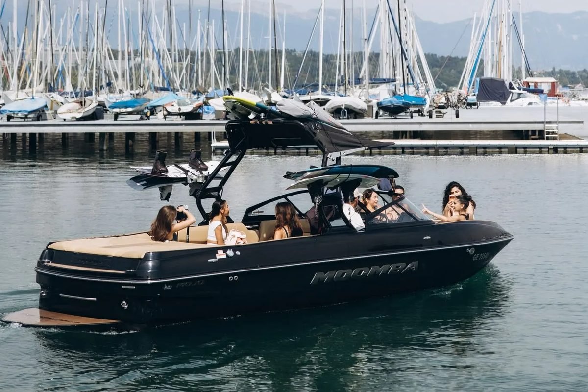 O barco preto da Evasion Wake-Surf a sair da marina com um grupo a bordo, mastros de veleiros ao fundo.