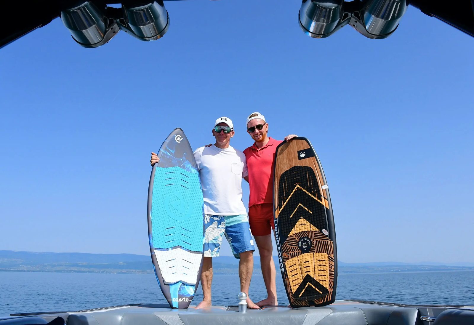 Dois membros da equipa de pé no barco, cada um com a sua prancha de wakesurf, contra o céu azul.