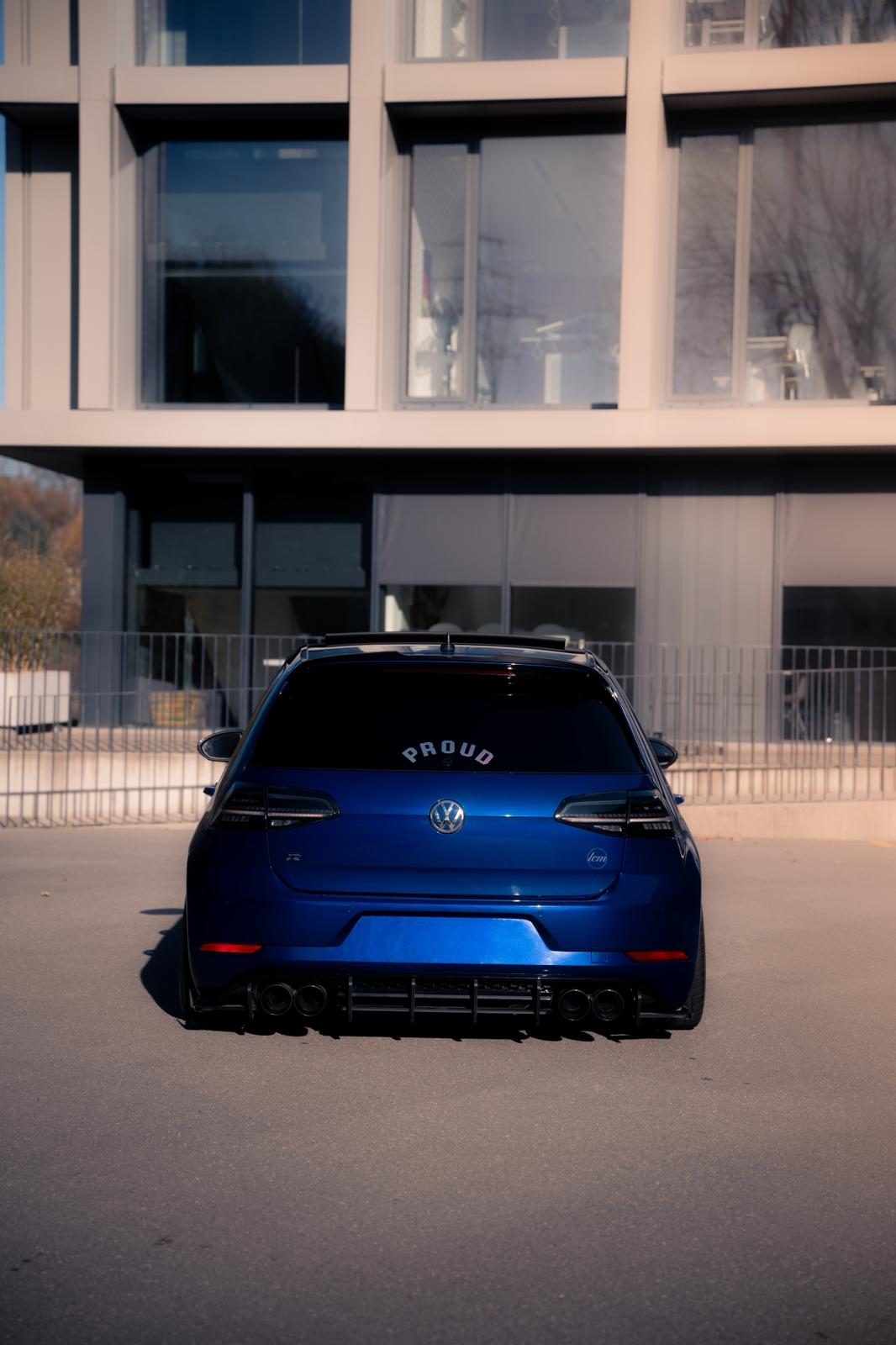 Um Volkswagen Golf azul rebaixado, visto de trás, estacionado em frente a um edifício de escritórios com fachada de vidro.