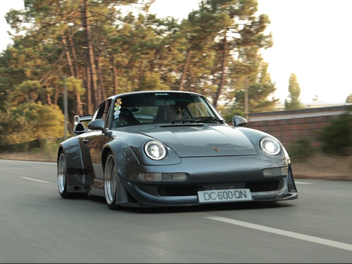 Um Porsche 911 cinzento de guarda-lamas largos em andamento numa estrada ladeada de pinheiros.